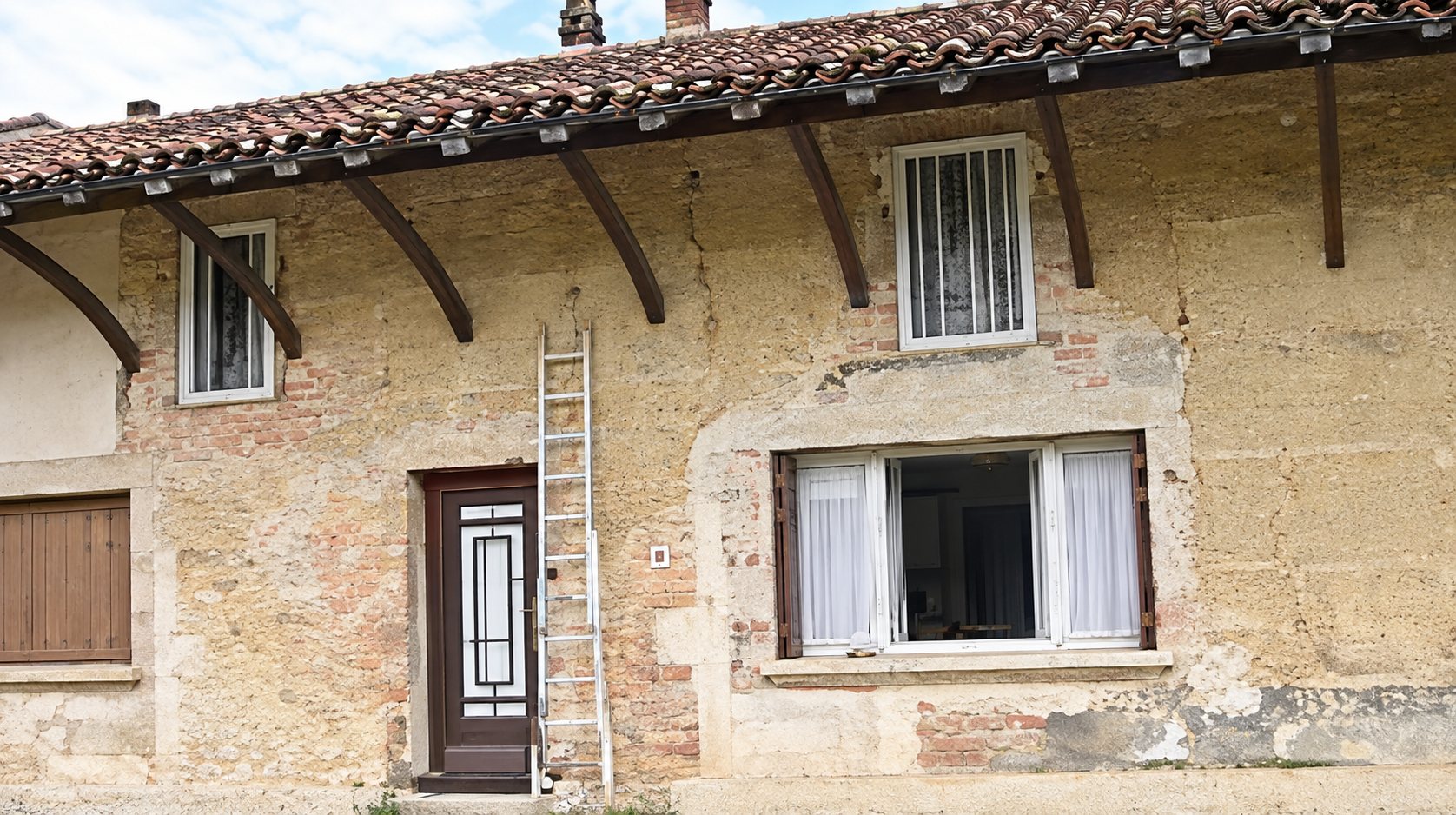 Façade d'une maison ancienne en pierres apparentes et enduit abîmé, avec des fenêtres à volets et une échelle appuyée près de la porte.