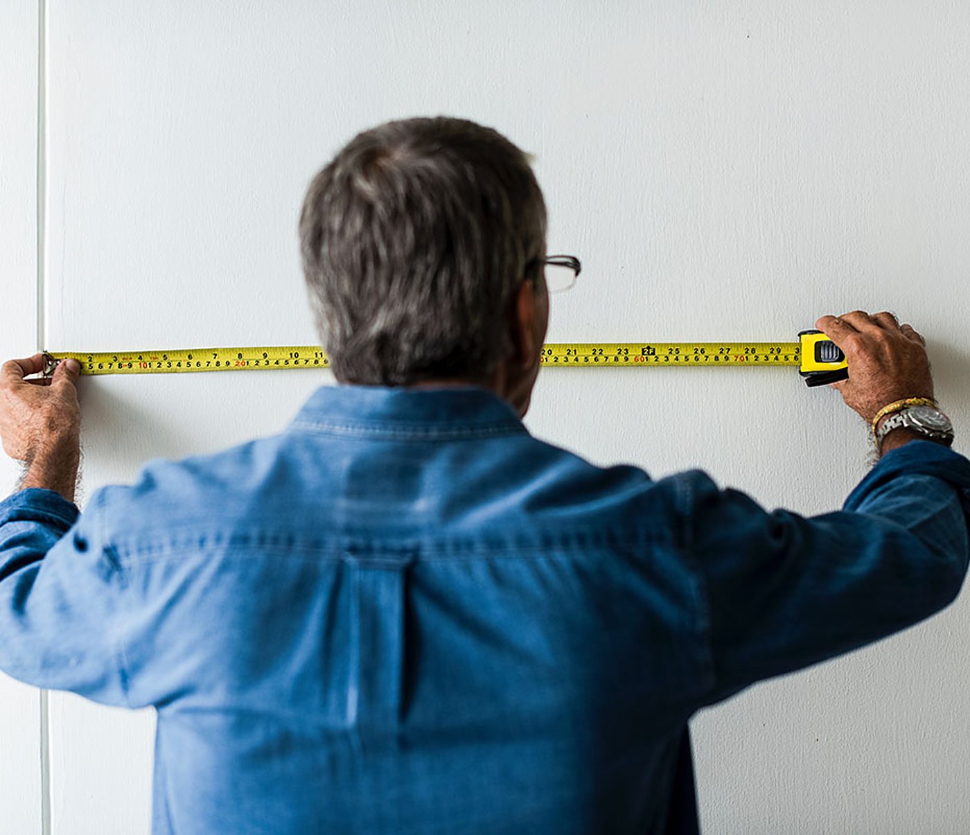 Un homme aux cheveux gris, vu de dos et vêtu d'une chemise en jean, tend un mètre ruban jaune contre un mur blanc pour prendre une mesure.